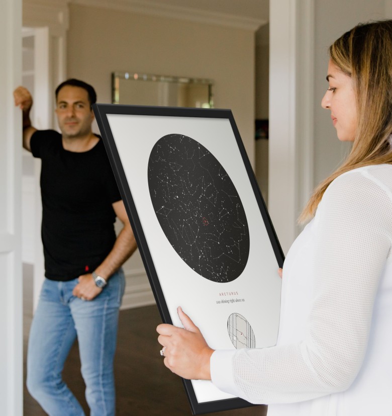 A woman holds a star map poster, the Our star design, in a black frame, a man watches from the doorway