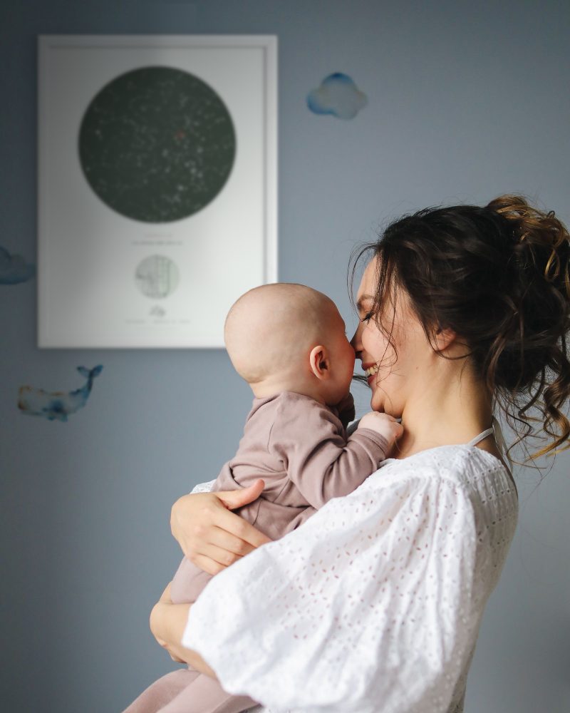 A mother holds her baby in a nursery, a star map poster in a white frame on the wall
