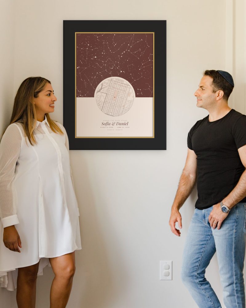 A couple stands by a star map poster, the Under the same stars design, in a black and gold frame
